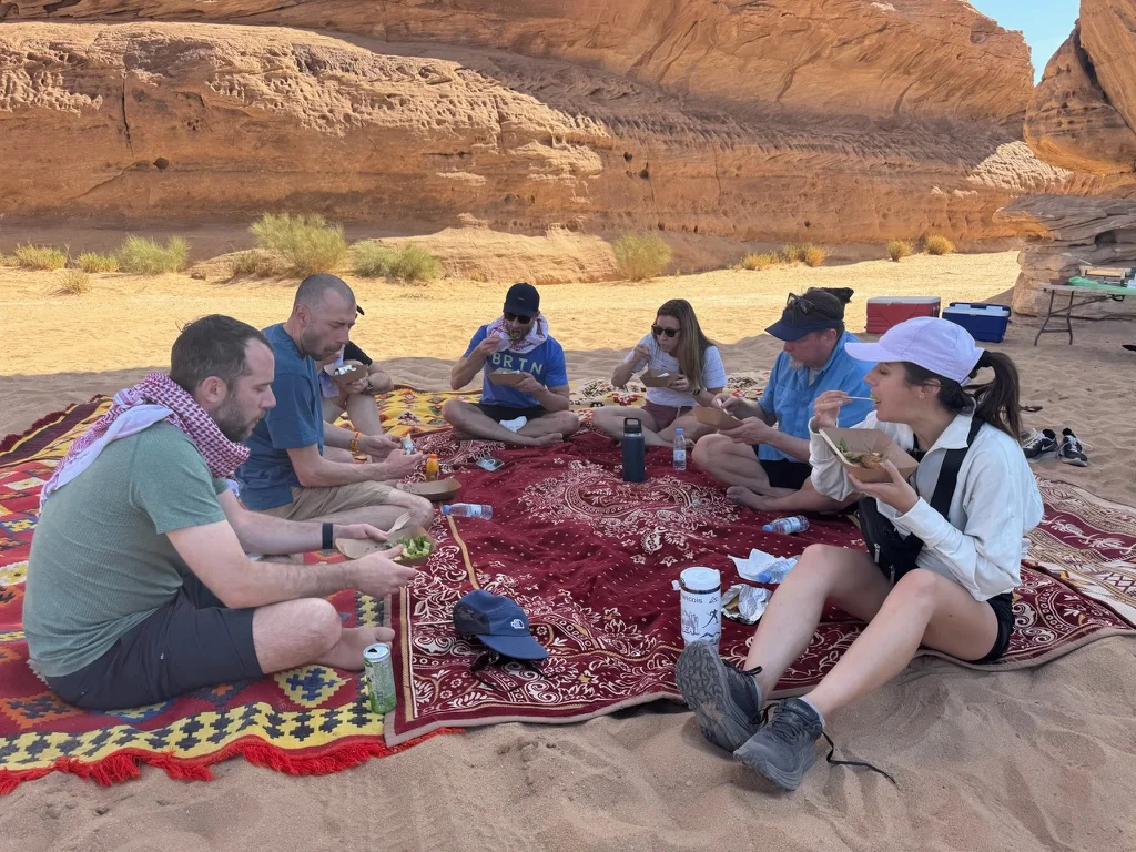 An executive team sharing a meal on a Bedouin rug among the red rock of AlUla, Saudi Arabia